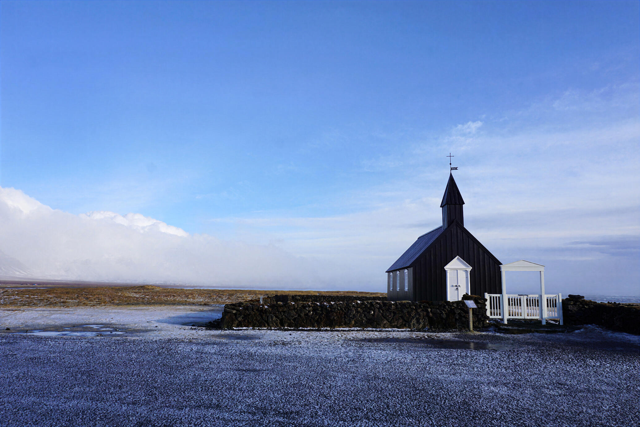 A quiet winter scene of Búðakirkja, Iceland’s iconic Black Church, standing alone against a vast blue sky and snowy landscape on the Snæfellsnes Peninsula. The stark contrast between the dark church, pale ground and distant mountains creates a serene and t