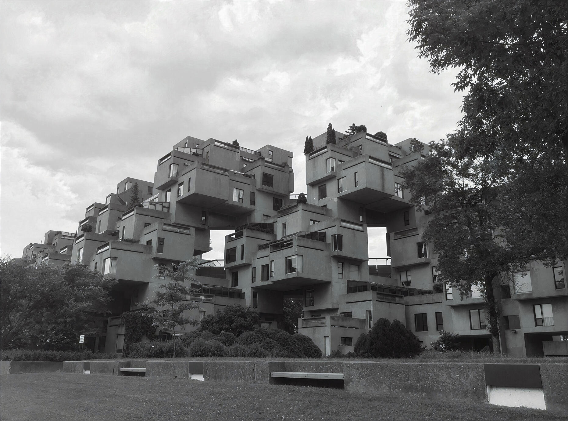 A striking black and white photograph of Montréal's distinctive Habitat 67, where geometric concrete homes rise in an intricate arrangement of stacked forms. The dramatic architecture, surrounding greenery and open sky create a timeless study of modern urb