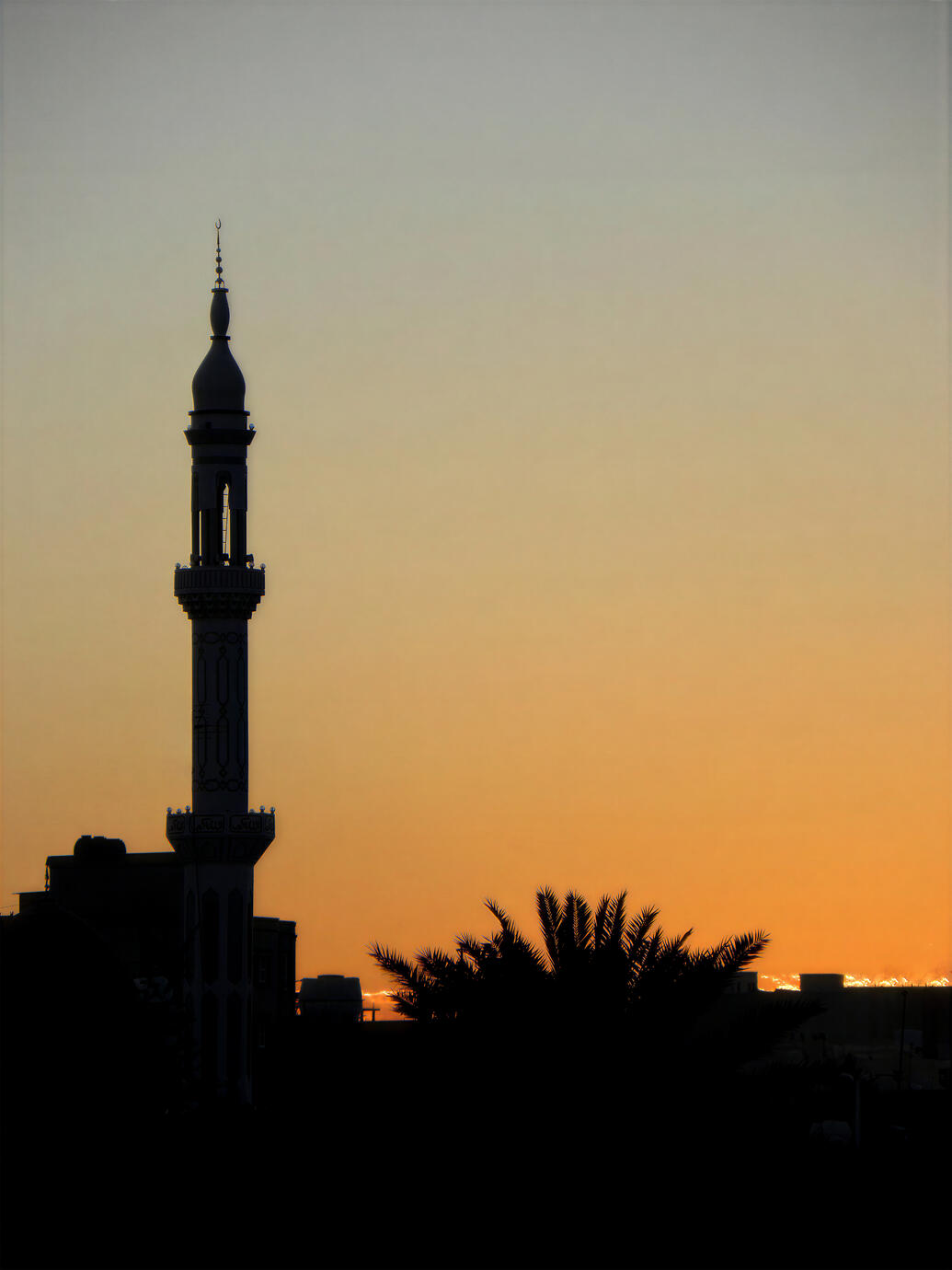 A dramatic sunset photograph of a traditional mosque minaret silhouetted against the golden sky in Salakh, Qeshm Island, Iran. The dark architectural silhouette, palm trees and glowing horizon capture the quiet character of southern Iran and the Persian Gu