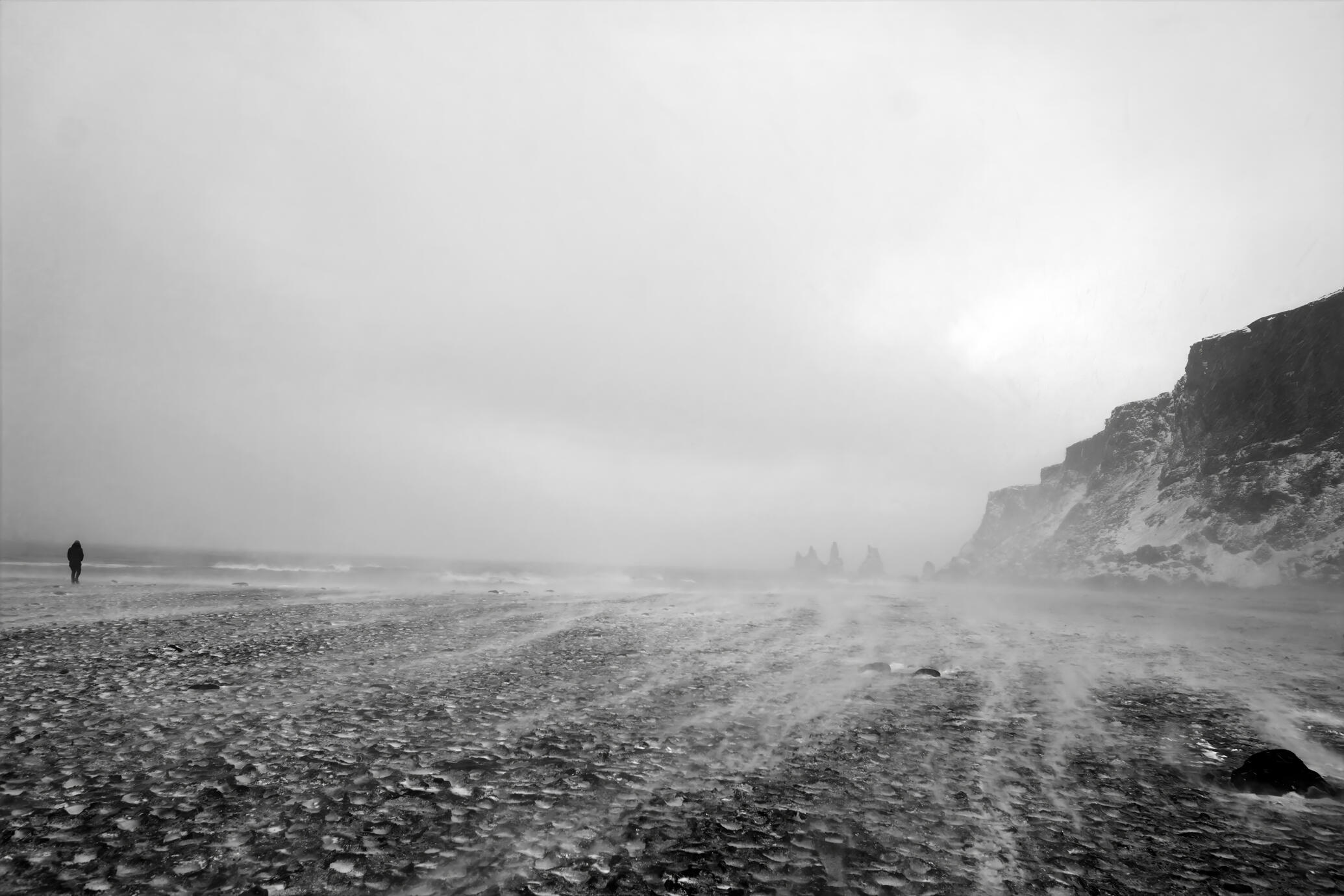 A moody black and white photograph of an Icelandic coastline during a powerful coastal storm. Wind drives mist and spray across the dark pebble beach as a solitary figure walks toward the cliffs in the distance. The stark landscape, soft light and atmosphe