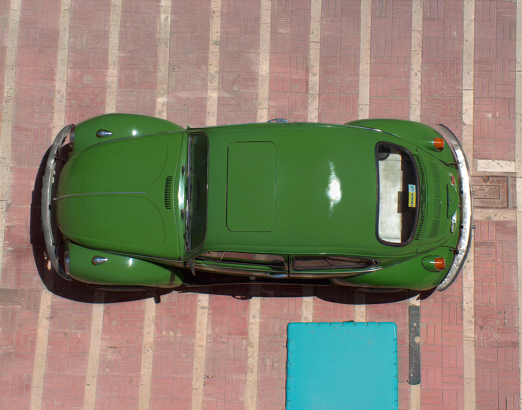 A striking overhead photograph of a classic green Volkswagen Beetle parked on the brick streets of Tehran, Iran. The bold green body, geometric pavement and contrasting turquoise detail create a distinctive vintage composition.