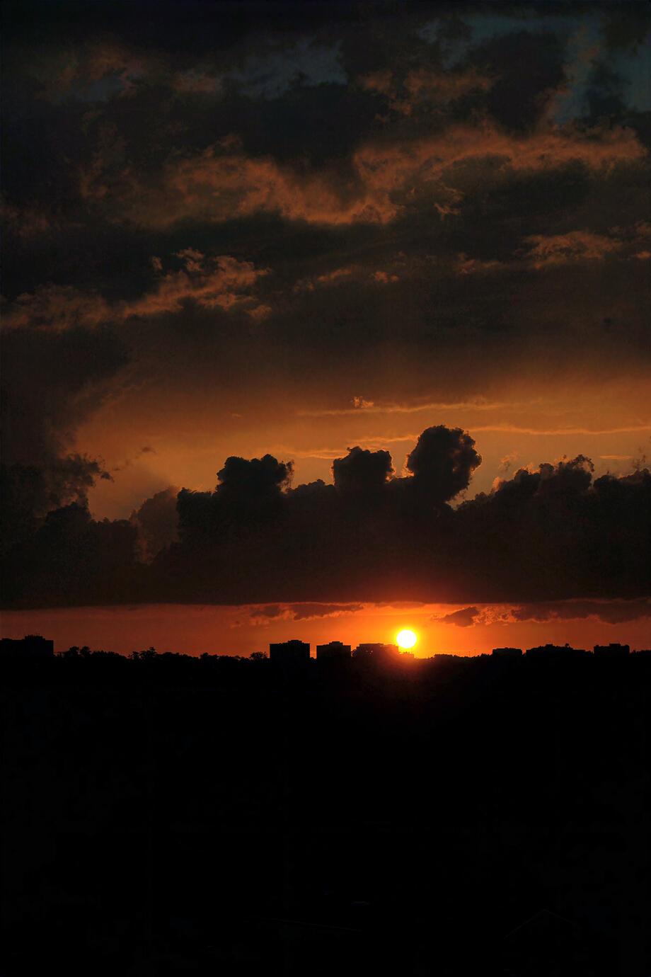 A dramatic Toronto sunset beneath towering clouds, with the city skyline silhouetted against a glowing amber horizon. The deep shadows and fiery light create a powerful, atmospheric evening scene.