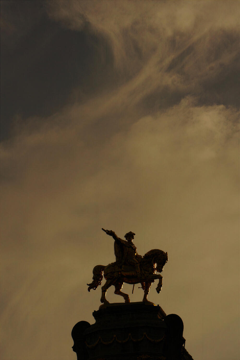 A dramatic silhouette of the equestrian statue of Godfrey of Bouillon beneath a vast, cloud-filled sky in Brussels. Warm golden light and deep shadows give the historic monument a timeless and almost cinematic presence.